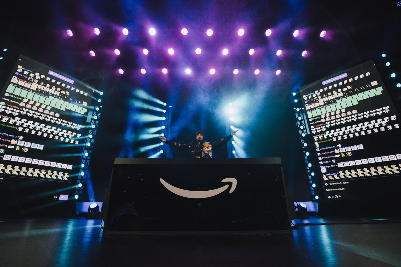 Live broadcast stage: a DJ at center between two towering chat-feed LED walls, beneath a matrix of magenta and blue beam fixtures cutting through haze