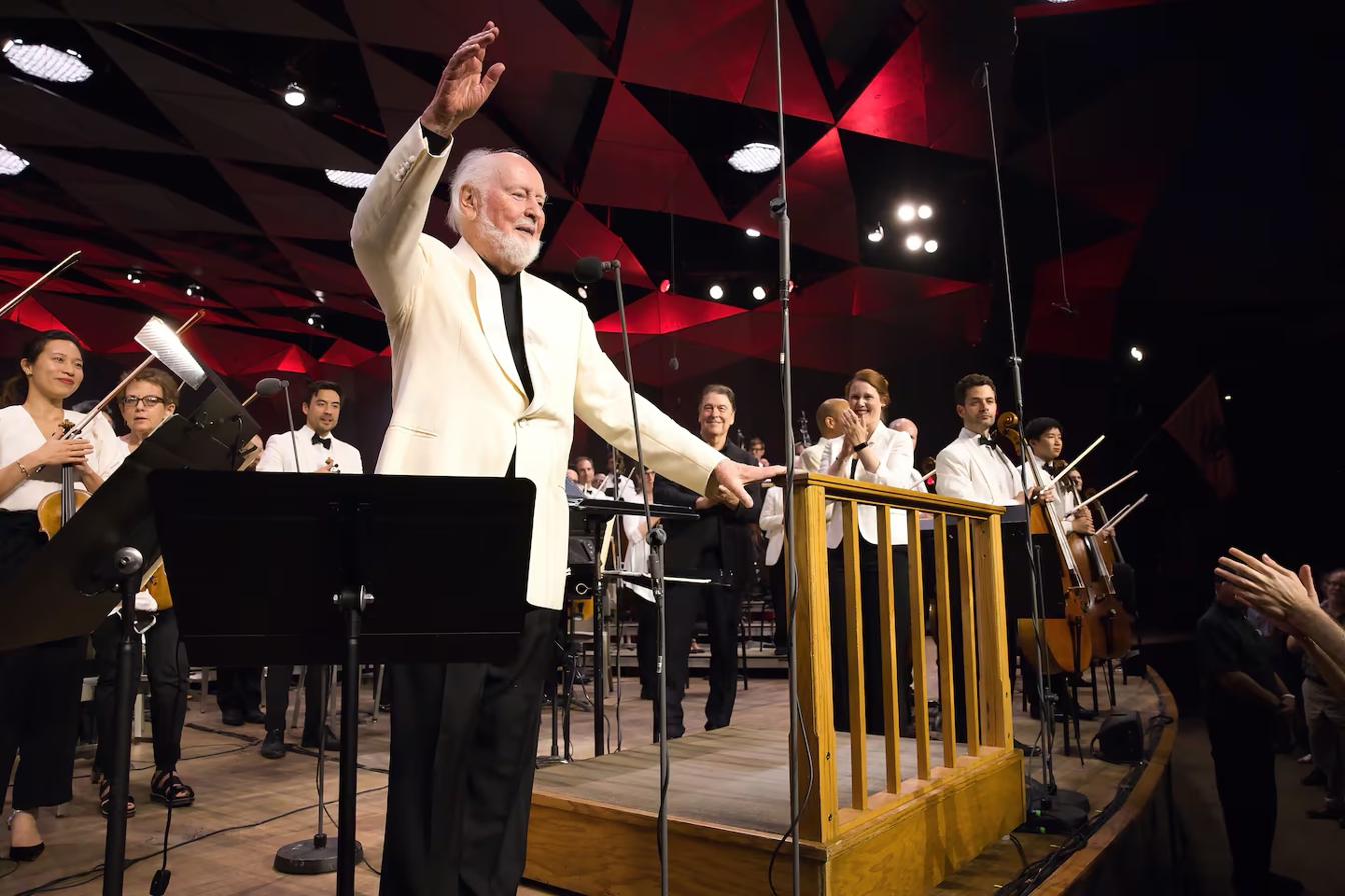A conductor in a white jacket acknowledging applause before an orchestra, beneath a faceted ceiling washed in deep red light