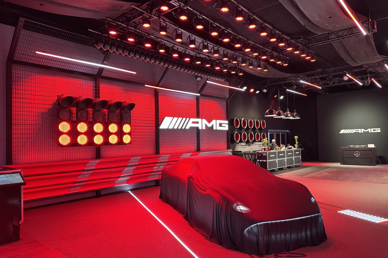 A premium automotive reveal space bathed in red light: a car under a black drape on red carpet, an illuminated AMG sign on a mesh wall, and a dense overhead grid of warm-toned fixtures