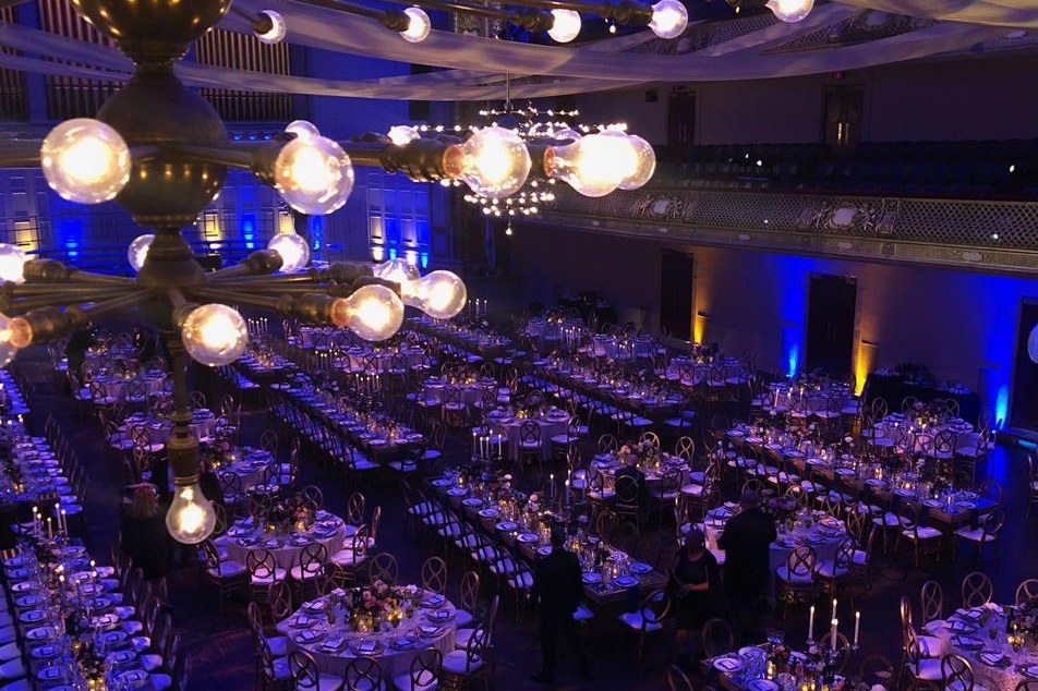 Historic concert hall transformed for a gala dinner: long candlelit tables under sapphire-blue uplighting, viewed from a balcony past brass chandeliers